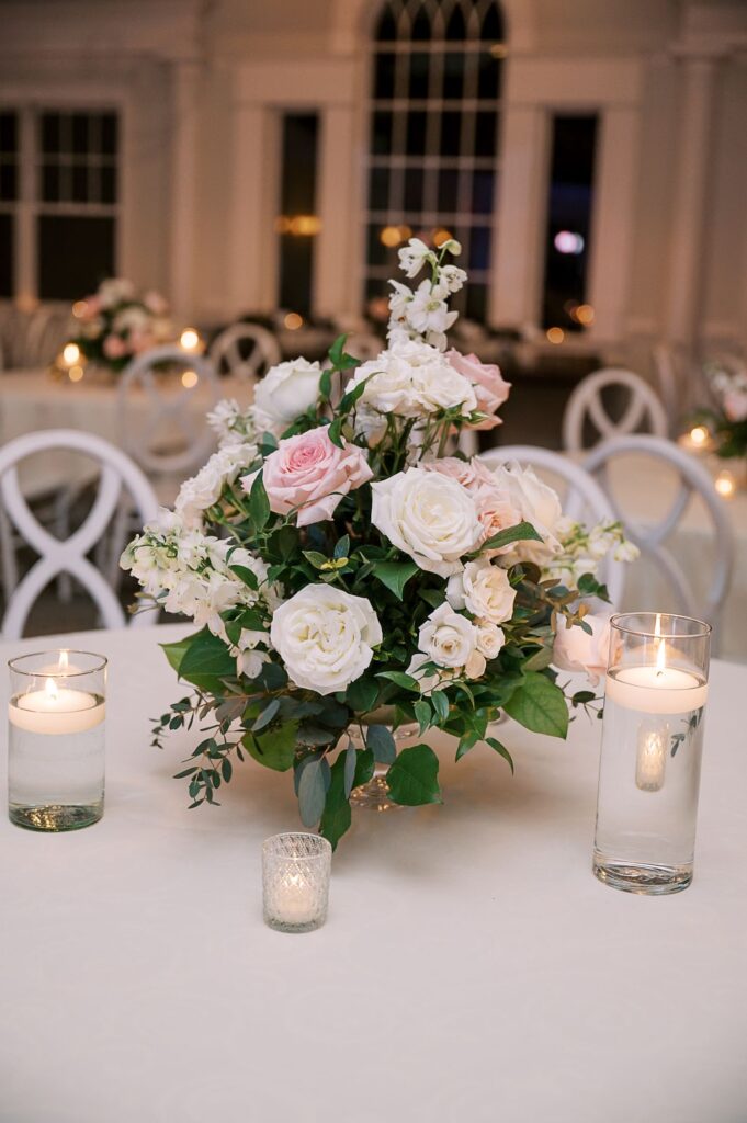 A floral centerpiece with white and pale pink roses sits on a white table, flanked by floating candle vases and a votive candle, in an elegant, softly lit room—perfect for a progressive buffet wedding in Atlanta.