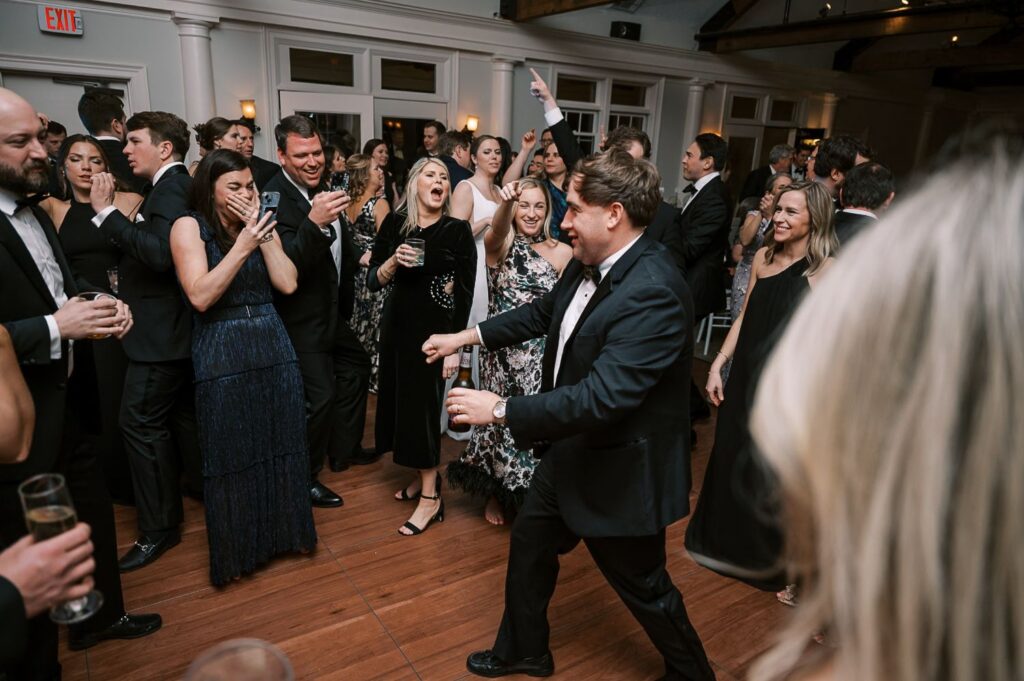 At a progressive buffet wedding in Atlanta, a man in a tuxedo dances energetically on a wooden floor, surrounded by smiling and laughing guests in formal attire. Some people are filming the lively celebration with their phones.