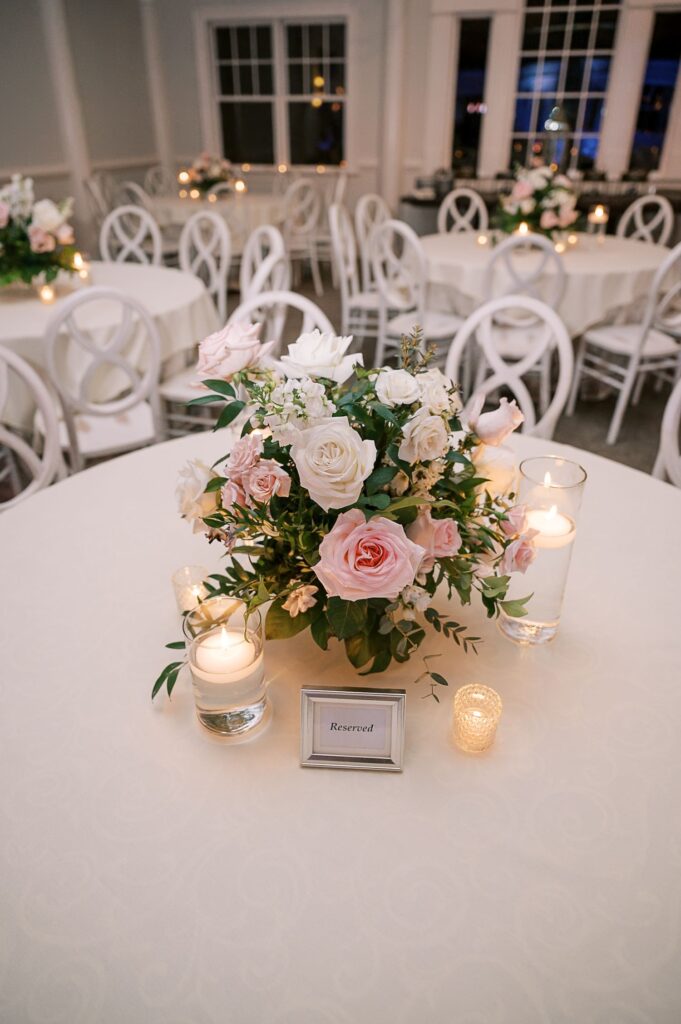 A round table with a floral centerpiece of white and blush roses, surrounded by lit candles and a framed “Reserved” sign, set for a progressive buffet wedding in Atlanta’s elegant event space with chic white chairs and decor.