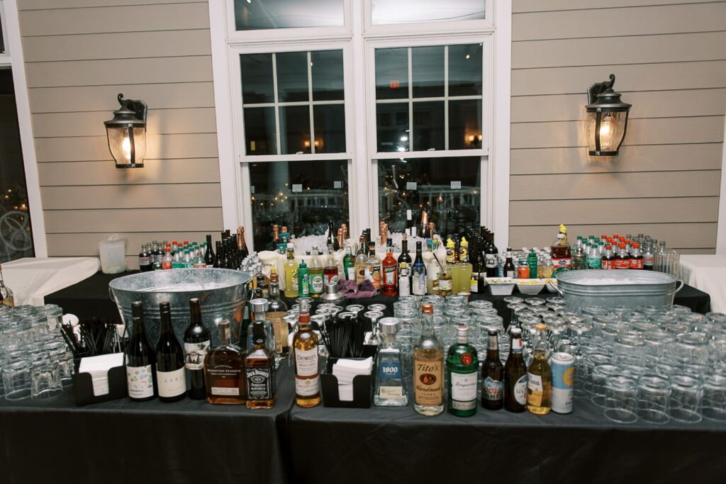 A large bar setup indoors with assorted liquor bottles, wine, mixers, and glassware on black tablecloth-covered tables in front of a window—perfect for a progressive buffet wedding in Atlanta—lit by two wall-mounted lantern lights.