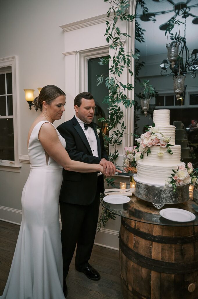 A bride and groom in formal attire stand together cutting a white, three-tiered wedding cake adorned with pink roses atop a wooden barrel, during their elegantly decorated progressive buffet wedding in Atlanta.
