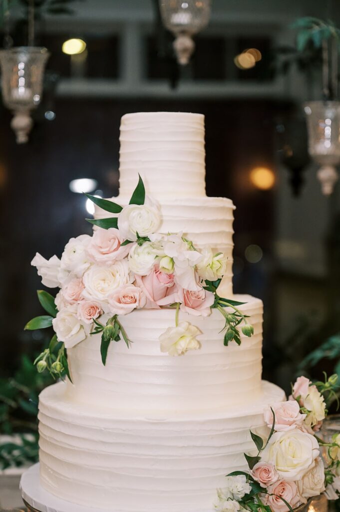 Three-tiered white wedding cake with textured icing, decorated with white and pale pink roses and greenery, set indoors with soft lighting and hanging glass lanterns—perfect for a progressive buffet wedding in Atlanta.