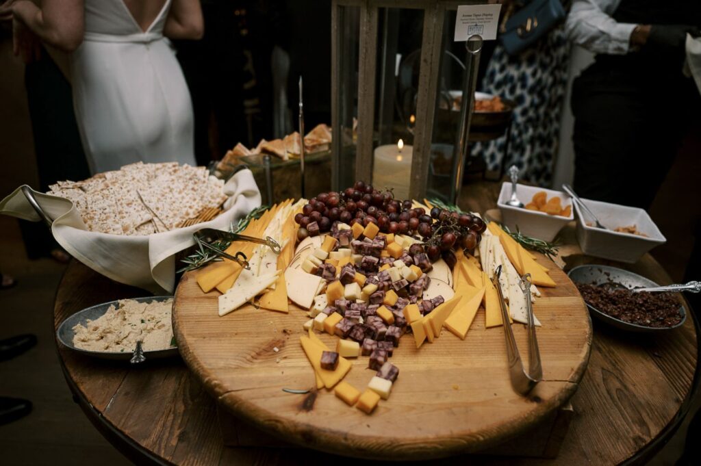 A round wooden platter topped with assorted cheeses, cubed meats, and red grapes, surrounded by crackers, spreads, and dried fruit on a rustic table at a progressive buffet wedding in Atlanta. People are visible in the background.