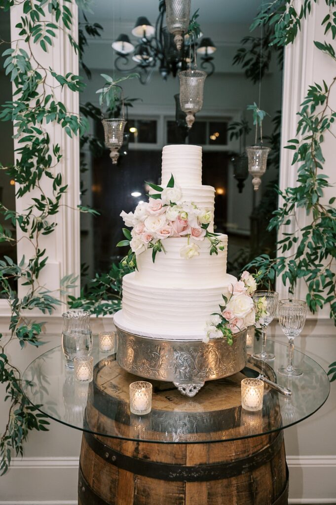 A three-tier white wedding cake decorated with pink and white flowers sits on a silver stand atop a wine barrel, part of a progressive buffet wedding in Atlanta. Candles, glassware, greenery, and hanging lights create an enchanting backdrop.