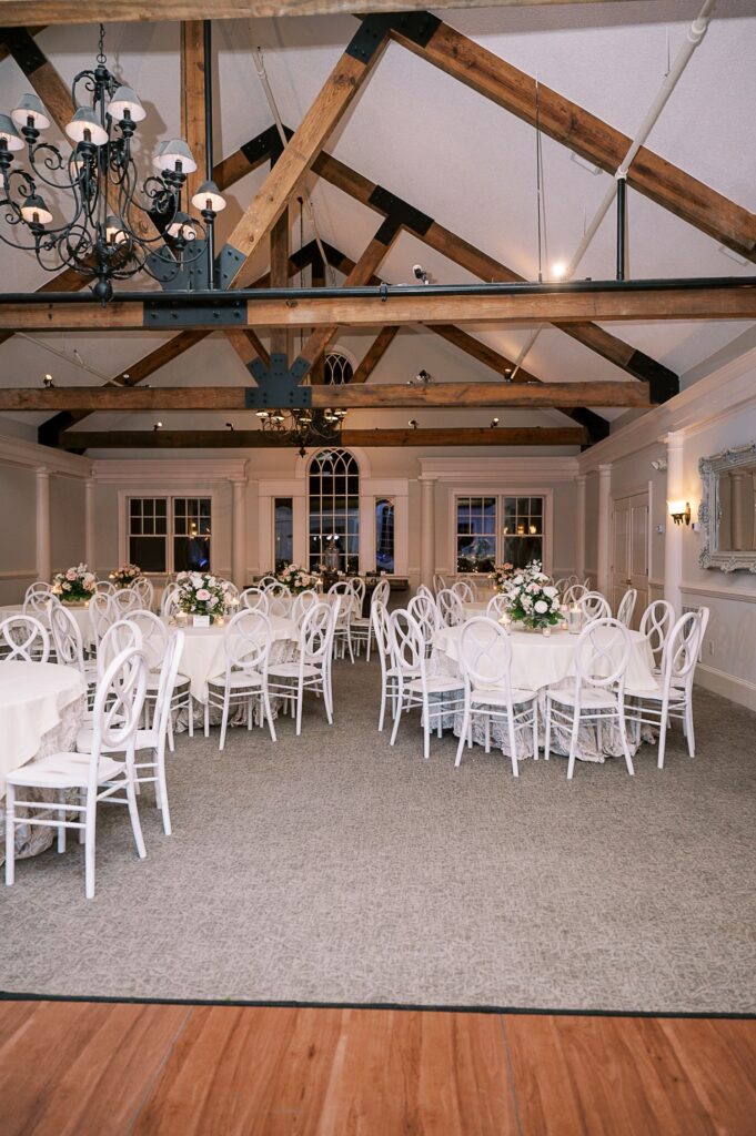 A decorated banquet hall in Atlanta hosts a progressive buffet wedding, with round tables draped in white tablecloths and floral centerpieces. White chairs, exposed wooden beams, and chandeliers add elegance to the bright, spacious room.