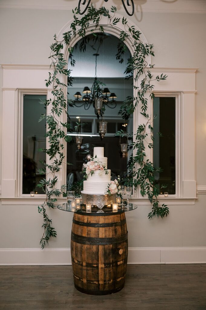 A white, three-tiered wedding cake adorned with pink and white flowers sits on a glass table atop a wooden barrel, surrounded by candles and greenery—perfect for a progressive buffet wedding in Atlanta, framed by tall windows and an arched mirror.