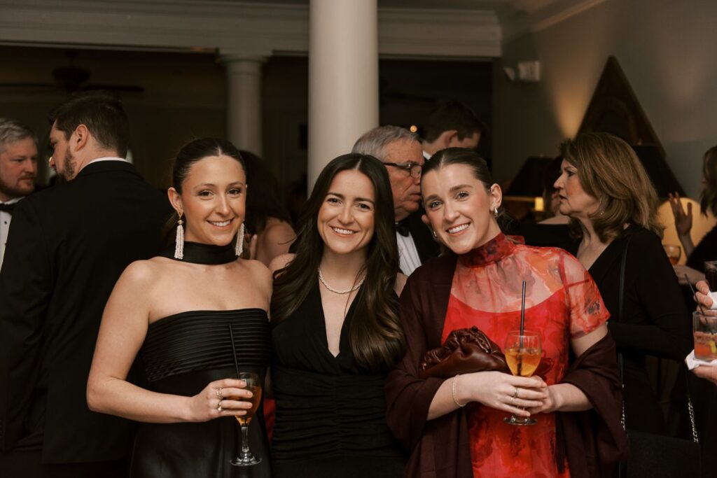 Three women dressed in formal attire smile and pose together at an indoor event in Atlanta. Holding drinks, they enjoy a progressive buffet wedding while other guests mingle in the lively background.