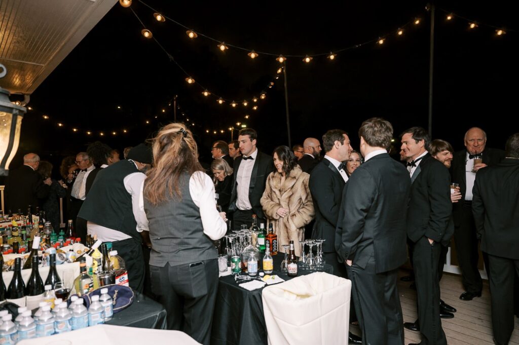 A group of people dressed in formal attire gather around a bar at an outdoor evening progressive buffet wedding in Atlanta, with string lights overhead and a bartender preparing drinks. Bottles and glasses are arranged on the bar.
