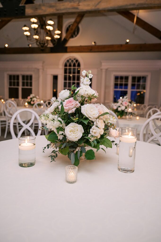 At a progressive buffet wedding in Atlanta, a round table with a white tablecloth features a floral centerpiece of white and pale pink roses, surrounded by three lit candles in glass holders. White chairs and other decorated tables are visible in the background.