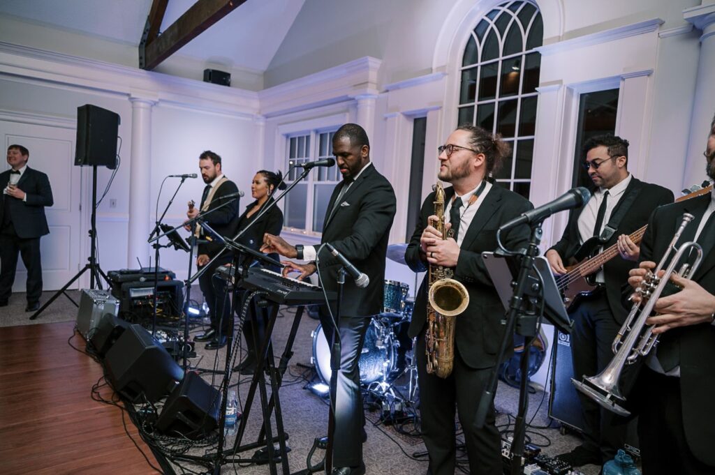 A band dressed in formal black attire performs indoors at a progressive buffet wedding in Atlanta; members play keyboard, saxophone, trumpet, bass guitar, and sing into microphones, with a man in a suit standing near a microphone in the background.