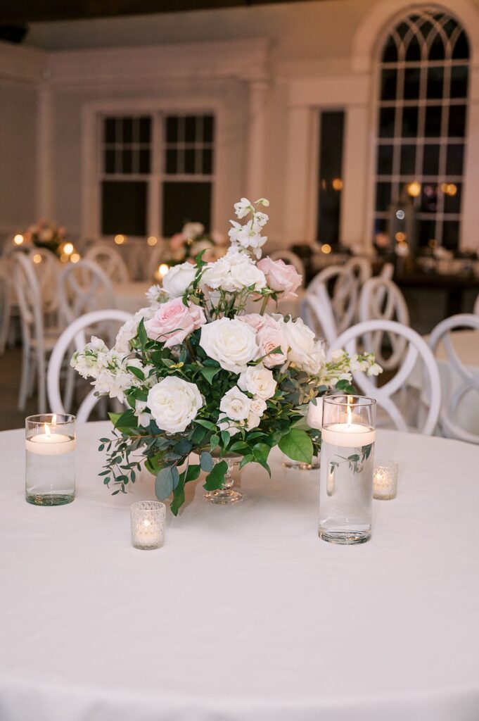 A round table with a white tablecloth features a centerpiece of white and pale pink flowers in a glass vase, surrounded by floating candles—perfect for an elegant, softly lit room at a progressive buffet wedding in Atlanta.