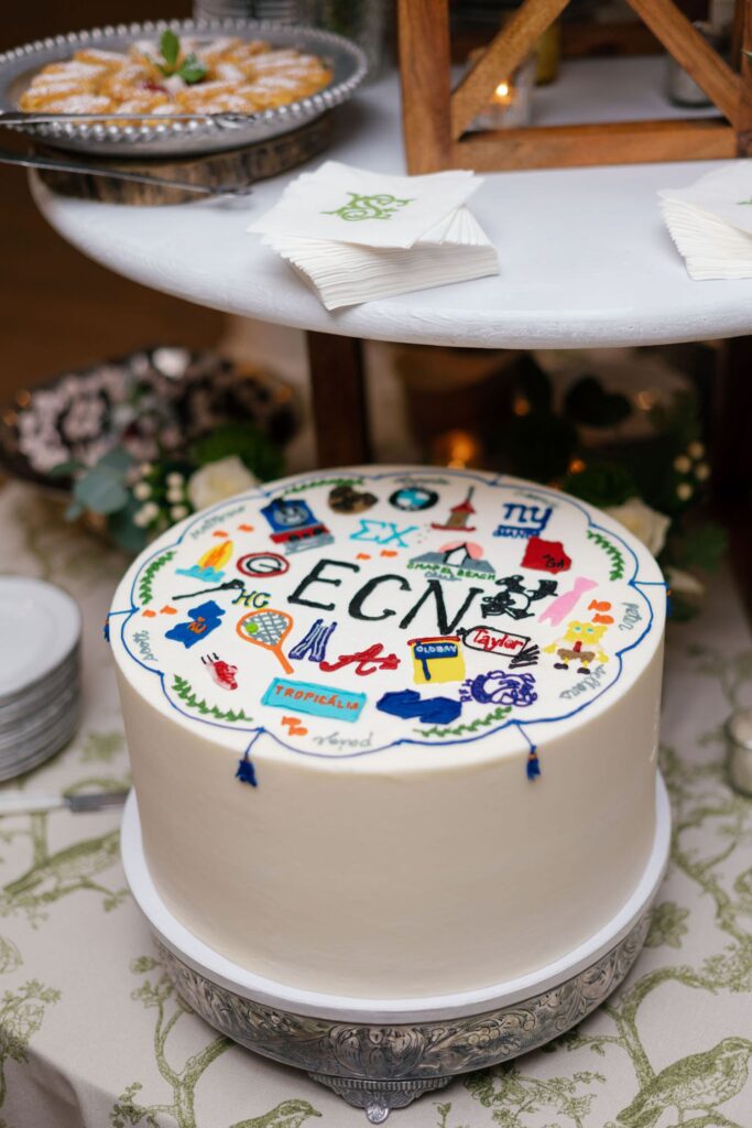 A round white cake decorated with colorful travel and hobby-themed designs and the letters EGN in the center, displayed on a silver stand—perfect for a Chastain Horse Park rehearsal dinner—beneath a marble tray with cookies and stacked napkins.