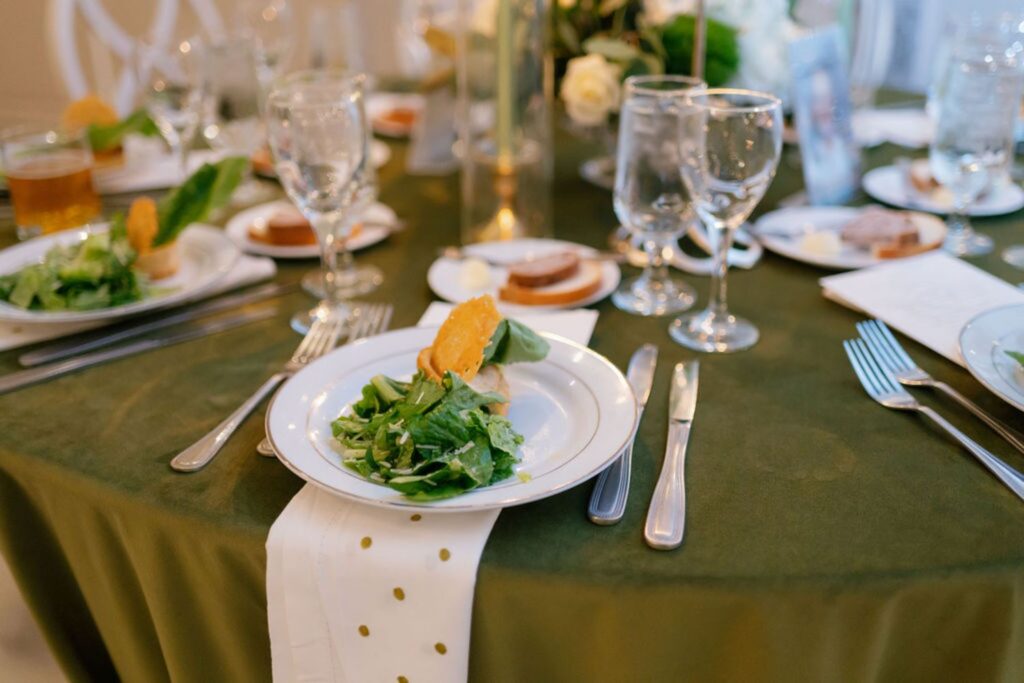 A round table set for a Chastain Horse Park rehearsal dinner features a green tablecloth, glassware, and white plates with leafy green salad and orange slices; white napkins with gold dots are neatly placed under the plates.