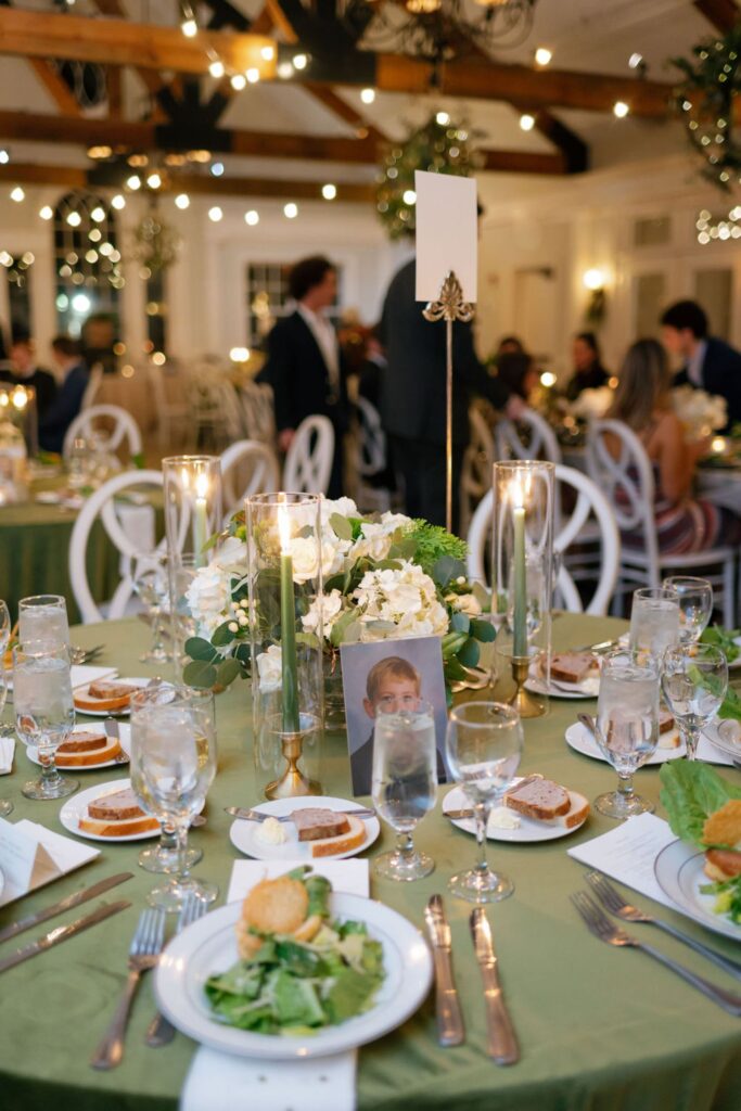A round table set for a formal Chastain Horse Park rehearsal dinner features a green tablecloth, white flowers, tall candles, plates of salad, bread, and water glasses. A child's photo is at the center; guests mingle in the softly lit background.