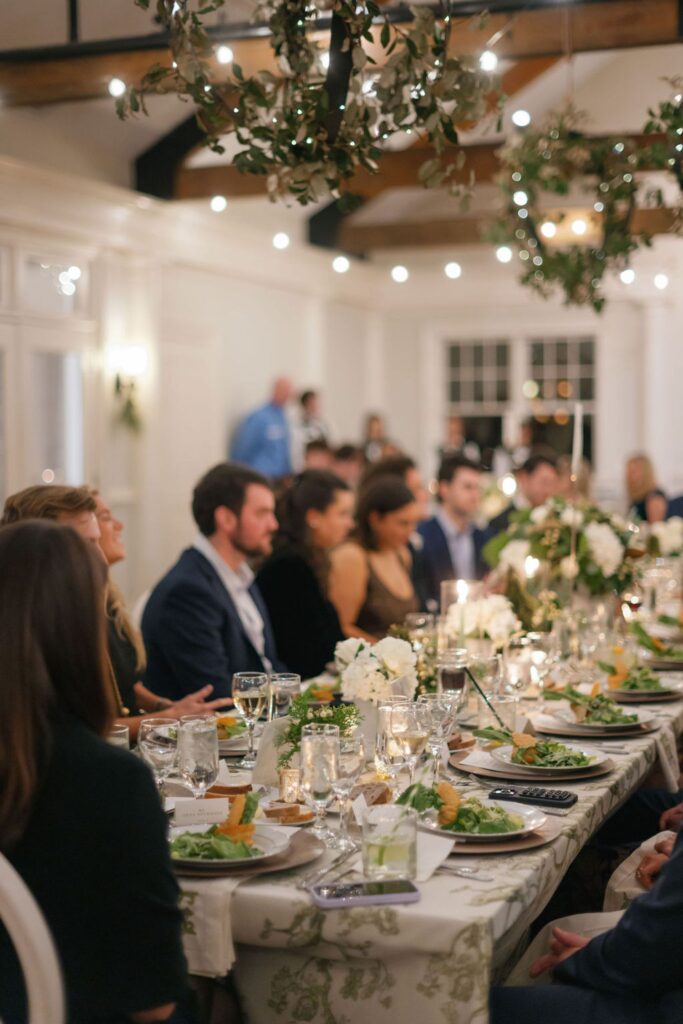 A group of people dressed formally sit around a long, elegant dining table decorated with white flowers, candles, and greenery, enjoying a meal in a warmly lit, festive indoor setting for a Chastain Horse Park rehearsal dinner.