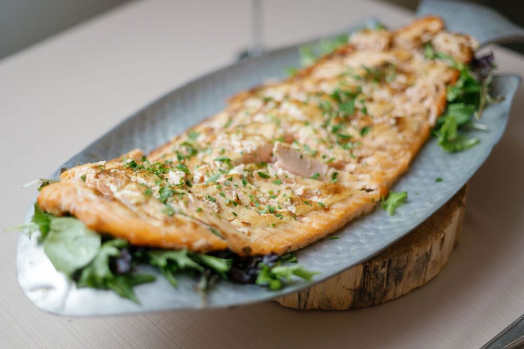 A whole grilled fish fillet garnished with chopped herbs is served on a fish-shaped metal platter lined with fresh greens, perfect for an elegant Chastain Horse Park rehearsal dinner and beautifully presented on a wooden board.