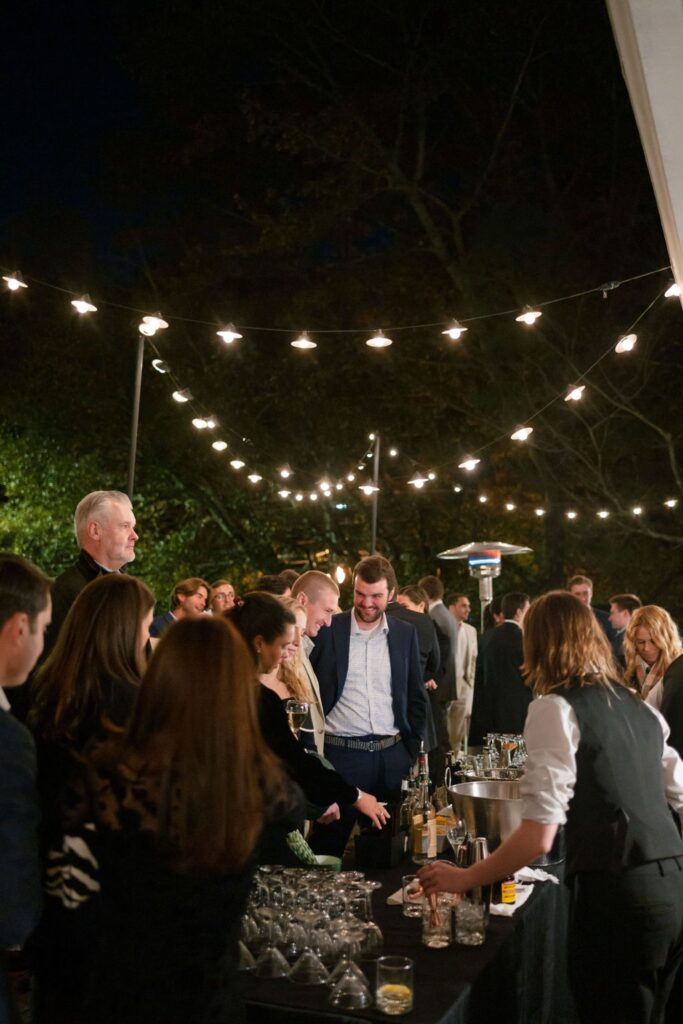 A group of people dressed in formal attire gather around an outdoor bar at night at a Chastain Horse Park rehearsal dinner, with string lights overhead creating a festive atmosphere as a bartender serves drinks and guests mingle.