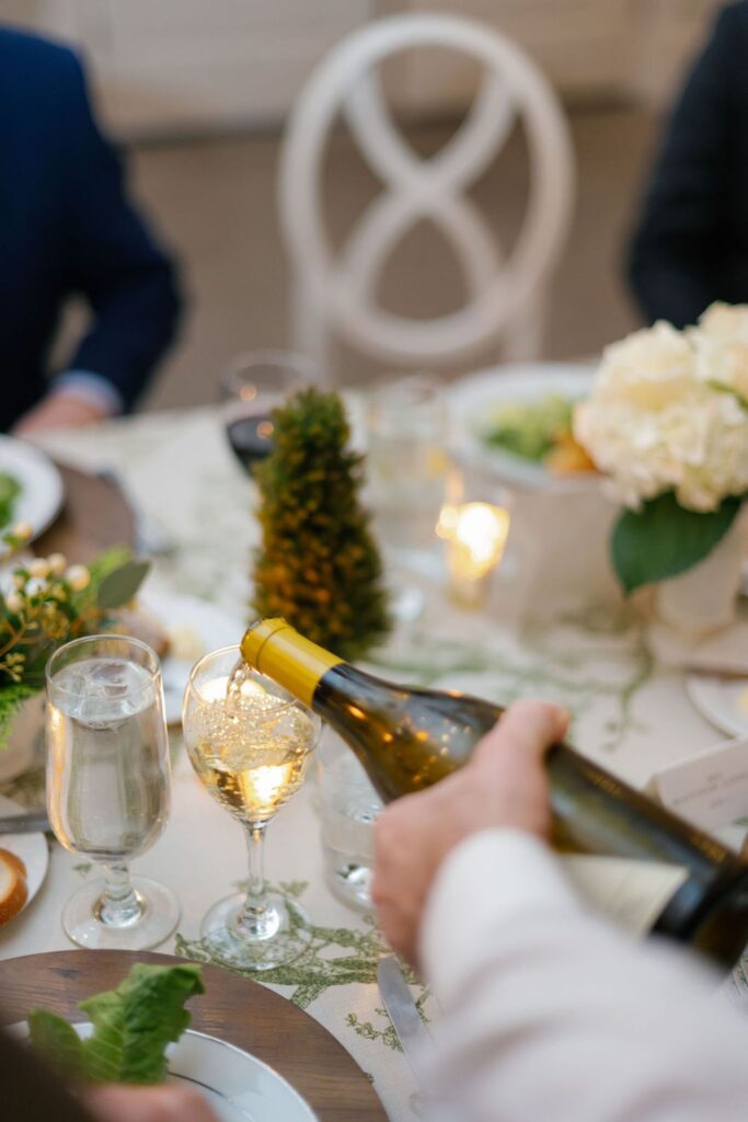 At a Chastain Horse Park rehearsal dinner, a person pours white wine into a glass at an elegantly set table adorned with flowers, a lit candle, water glasses, and plates of food, capturing a formal, festive dining atmosphere.