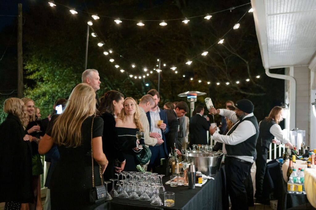 A group of people in formal attire gather at an outdoor evening Chastain Horse Park rehearsal dinner under string lights. A bartender pours drinks at a table stocked with glasses and bottles, while guests mingle and chat.
