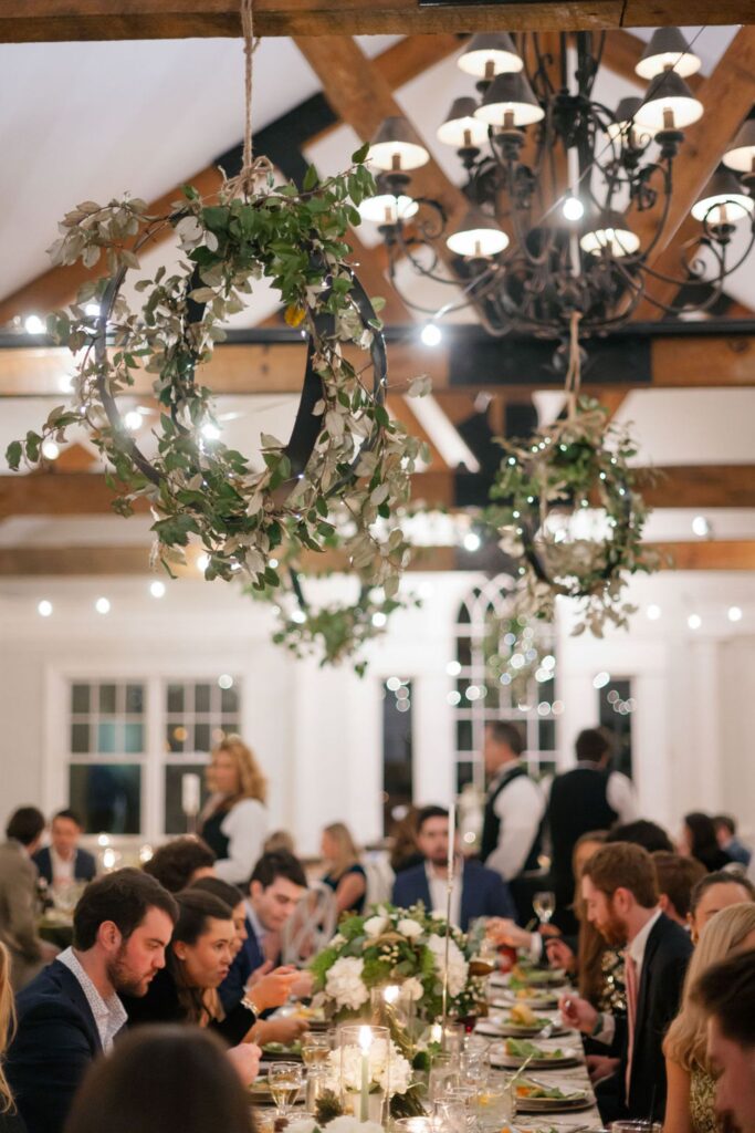 Guests gather at long, elegant tables set for a Chastain Horse Park rehearsal dinner in a warmly lit room with wooden beams. Hanging wreaths with greenery and string lights adorn the ceiling as servers stand in the background.