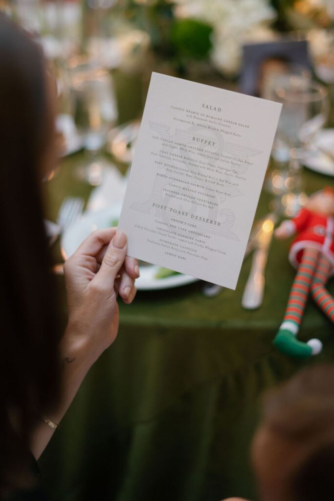 A person holds a dinner menu at a table set for a formal event, with plates, cutlery, glasses, and an elf doll decoration on a green tablecloth in the background at a Chastain Horse Park rehearsal dinner.