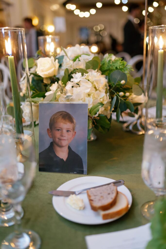 A framed school photo of a young boy is displayed on a table at the Chastain Horse Park rehearsal dinner, decorated with white flowers, green foliage, and tall lit candles. In the foreground, there is a plate with bread, butter, and a knife.