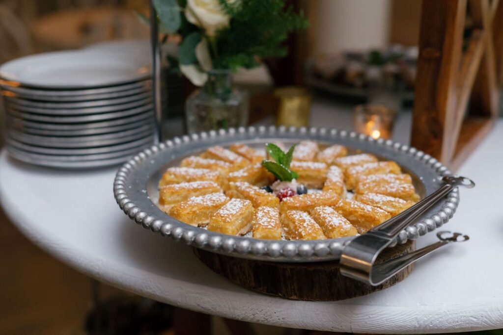 A round silver tray with pieces of pastry dusted with powdered sugar, arranged in a circle and garnished with mint, sits on a white table—perfect for a Chastain Horse Park rehearsal dinner—beside a stack of plates and cutlery.