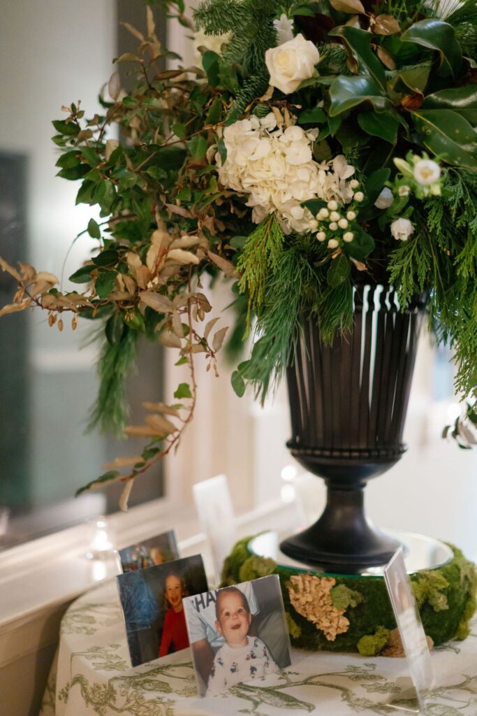 A large floral arrangement in a black vase sits on a table with a green patterned cloth, set for a Chastain Horse Park rehearsal dinner. Several framed photos, including baby and child portraits, are displayed around the base of the arrangement.