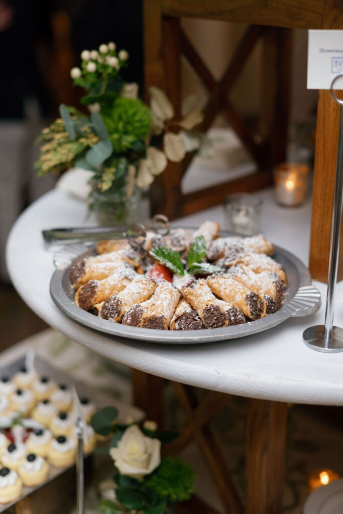 A silver tray with chocolate-dipped pastries dusted with powdered sugar, garnished with strawberries and mint, sits on a round white table at a Chastain Horse Park rehearsal dinner, surrounded by flowers and a glowing candle.