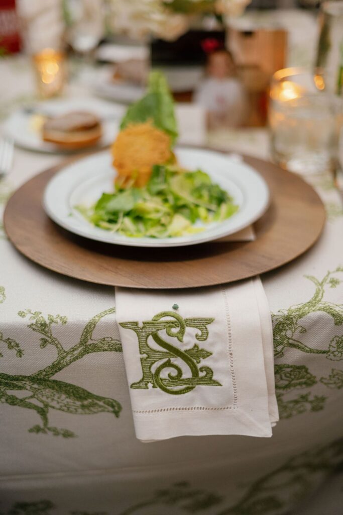 A white napkin with a green embroidered letter S rests on a patterned tablecloth at a Chastain Horse Park rehearsal dinner, next to a plate of salad topped with a Parmesan crisp and set on a wooden charger, with candles softly glowing in the background.
