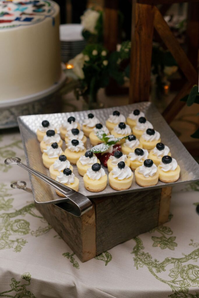 A tray of small dessert pastries topped with whipped cream and blueberries, displayed on a wooden stand with serving tongs nearby at a Chastain Horse Park rehearsal dinner. A decorated cake and greenery are visible in the background.