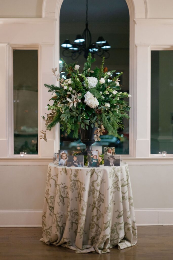 A large floral arrangement with white flowers and greenery sits on a round table draped with a patterned cloth. Several framed photos are arranged at the base, creating an elegant display for a Chastain Horse Park rehearsal dinner at night.