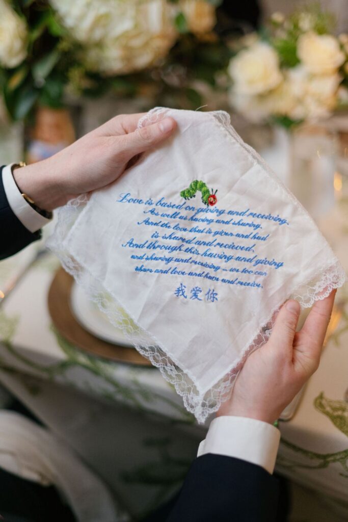 A person holds a lace-trimmed handkerchief embroidered with a poem, a green and red flower, and blue Chinese characters at a Chastain Horse Park rehearsal dinner. Elegant tableware and white flowers are visible in the blurred background.