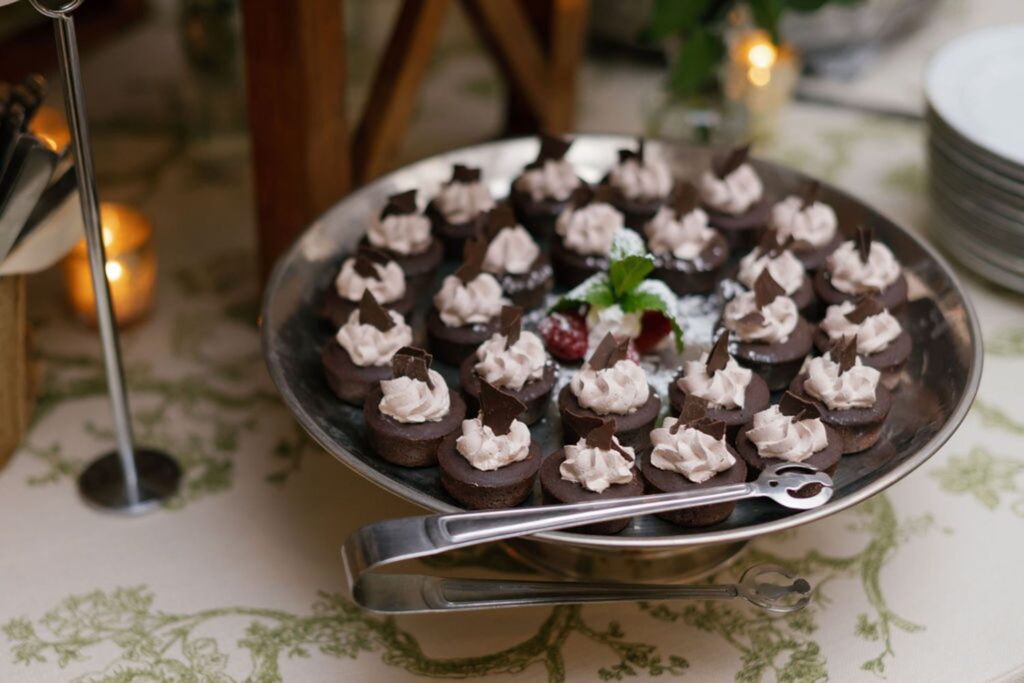 A metal tray filled with small chocolate desserts topped with whipped cream and chocolate pieces, garnished with mint and cherries, awaits guests at a Chastain Horse Park rehearsal dinner. Serving tongs, plates, and a candle are in the background.