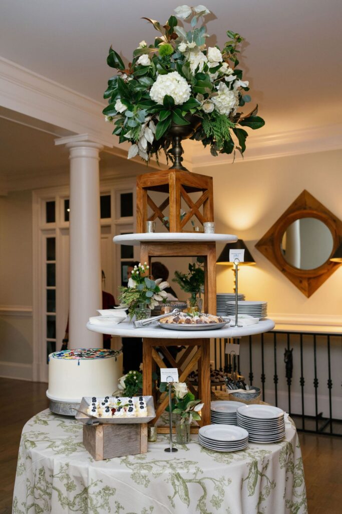 A tiered dessert display at a Chastain Horse Park rehearsal dinner features assorted sweets, a decorated cake, and floral arrangements on a round table with a white patterned cloth. Plates and utensils are neatly stacked nearby in a bright room.