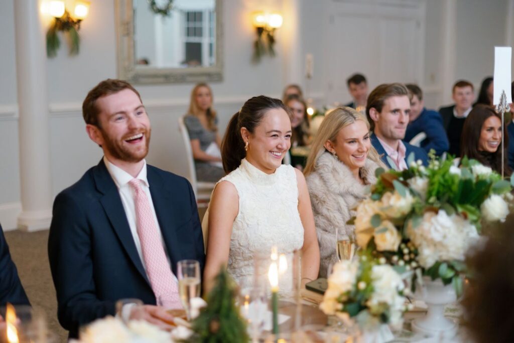 A group of well-dressed guests, smiling and laughing, sit at a table decorated with flowers and candles during a Chastain Horse Park rehearsal dinner in an elegant, softly lit room.