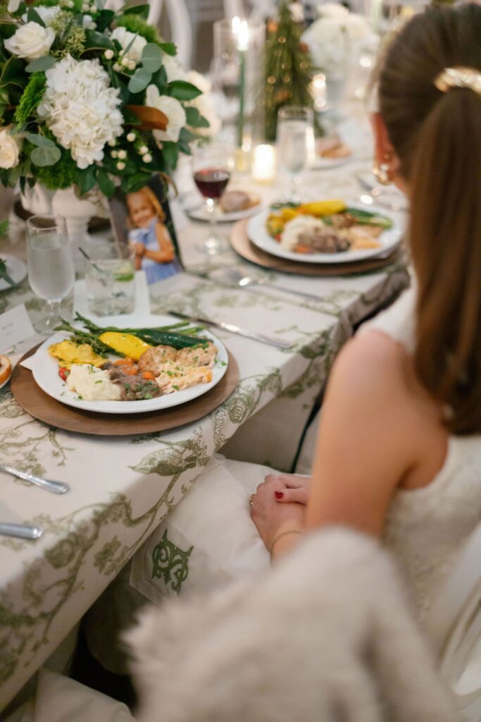 A woman in a white dress sits at a decorated table with a floral centerpiece, enjoying a plated meal of vegetables, rice, and meat during a Chastain Horse Park rehearsal dinner. The table is elegantly set with drinks, cutlery, and white linens.