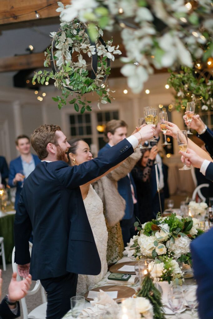 A bride and groom stand smiling and raise champagne glasses for a toast with guests at a beautifully decorated Chastain Horse Park rehearsal dinner, surrounded by flowers and hanging greenery.