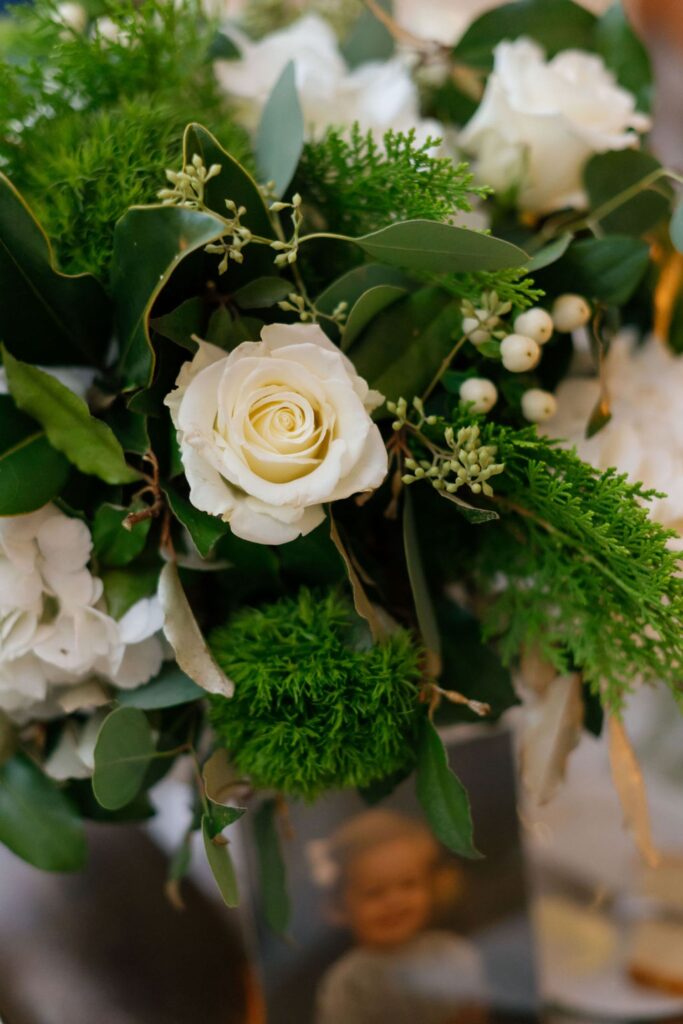 A close-up of a floral arrangement featuring white roses, green foliage, small white berries, and leaves at a Chastain Horse Park rehearsal dinner, with a blurred photo of a smiling child visible in the background.