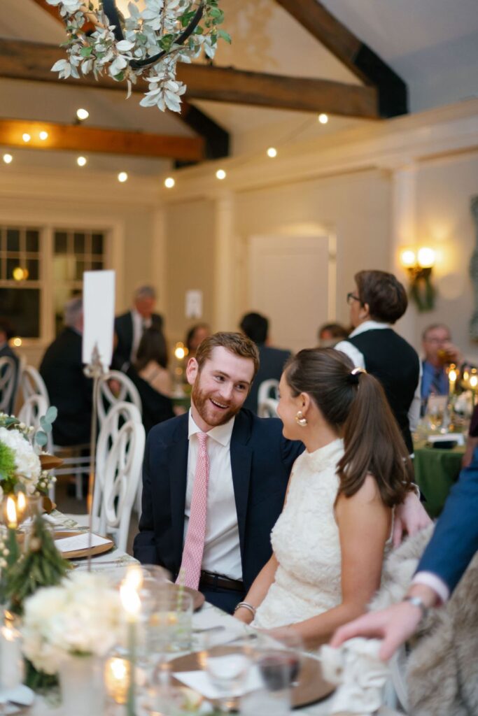 A man in a suit and a woman in a white dress sit closely at a decorated dinner table, smiling at each other, surrounded by other guests at an elegant, warmly lit Chastain Horse Park rehearsal dinner.