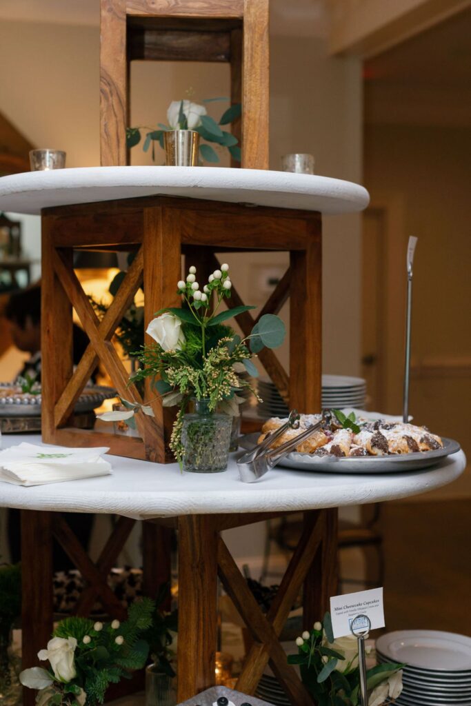 A tiered wooden display at the Chastain Horse Park rehearsal dinner holds plates, napkins, floral arrangements, and pastries dusted with powdered sugar. Small name cards and stacked white dishes sit on the round white shelves.