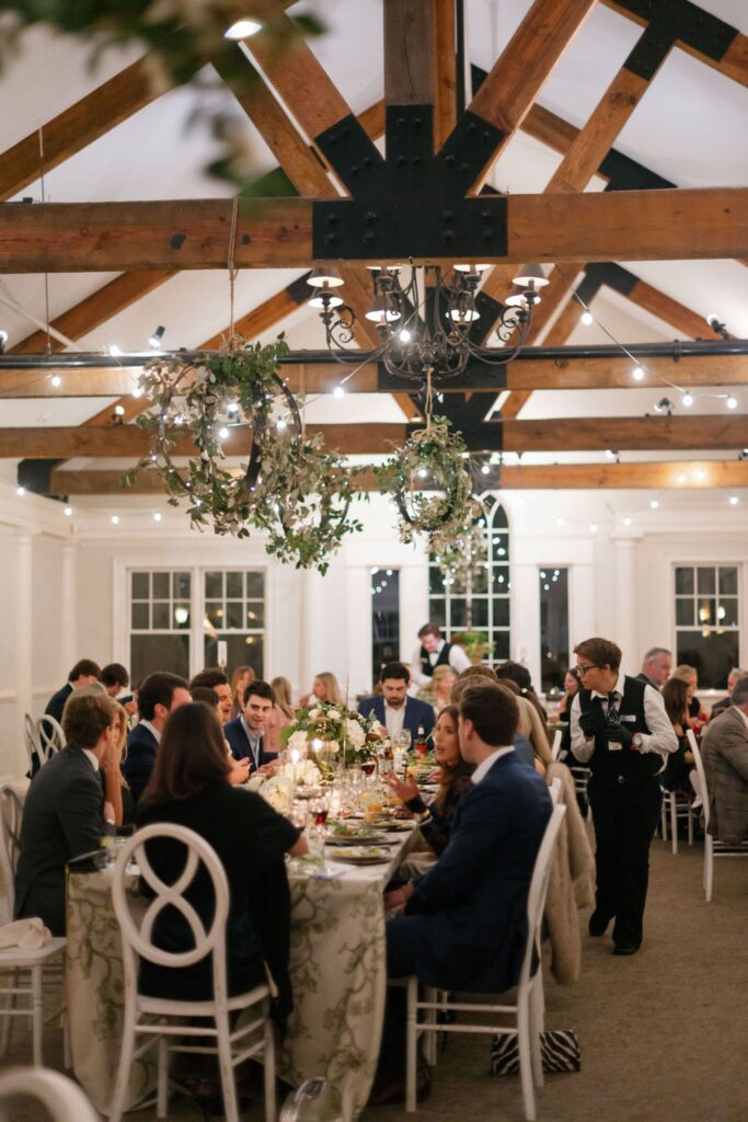 Guests sit at round tables with floral tablecloths under wooden beams and hanging greenery, enjoying a meal in a warmly lit, elegant indoor venue—an idyllic setting for a Chastain Horse Park rehearsal dinner. A server in a black vest attends to a table.