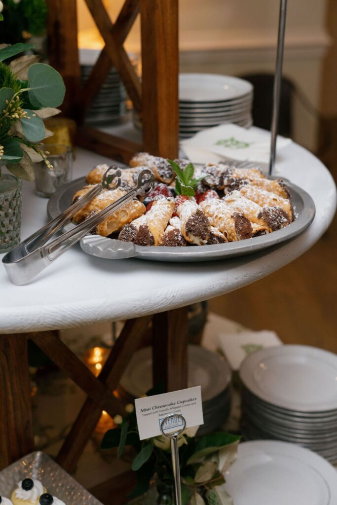 A platter of mini cheesecake cannolis dusted with powdered sugar sits on a white table at the Chastain Horse Park rehearsal dinner, with metal tongs, mint garnish, plates, and a sign labeled Mini Cheesecake Cannolis nearby.