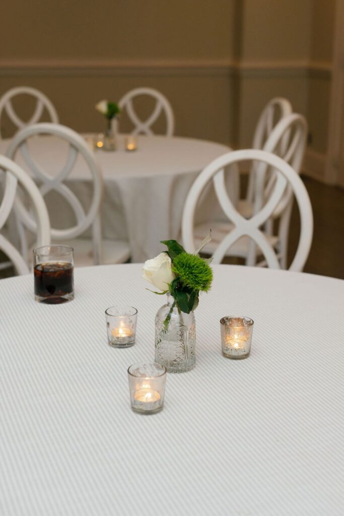 At a Chastain Horse Park rehearsal dinner, a round table with a white tablecloth displays a small vase with a white rose and greenery, three lit votive candles in glass holders, and a glass of dark soda. Similar tables and chairs are visible in the background.
