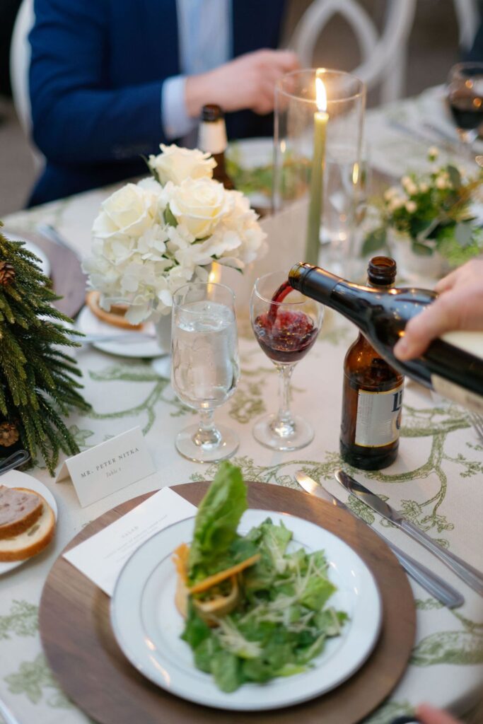 At a Chastain Horse Park rehearsal dinner, a hand pours red wine into a glass at a formal table set with salad, water, beer, white roses, candles, and a name card, as guests gather around the elegantly arranged scene.