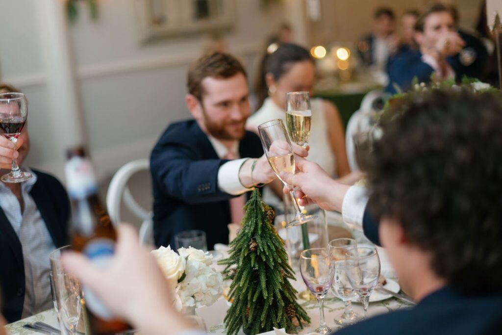 A group of people dressed formally sit around a table at a Chastain Horse Park rehearsal dinner, raising glasses in a toast. The table is decorated with a small pine tree centerpiece and white flowers, creating a festive and celebratory atmosphere.