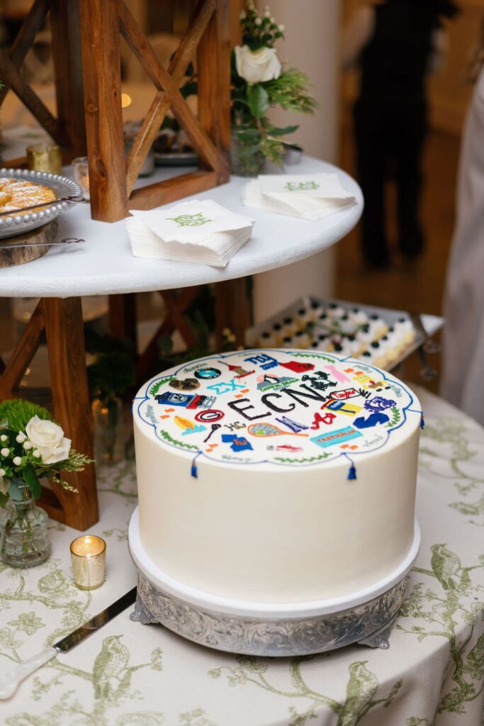 A round white cake decorated with colorful graduation-themed icons and the word ELECTRIC on top sits on a table at a Chastain Horse Park rehearsal dinner, surrounded by flowers, candles, napkins, and desserts in the background.