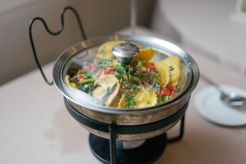 A stainless steel serving dish with yellow ravioli, cherry tomatoes, spinach, and herbs sits on a stand at a Chastain Horse Park rehearsal dinner; a spoon rests on top, and a white plate and spoon are visible in the background.