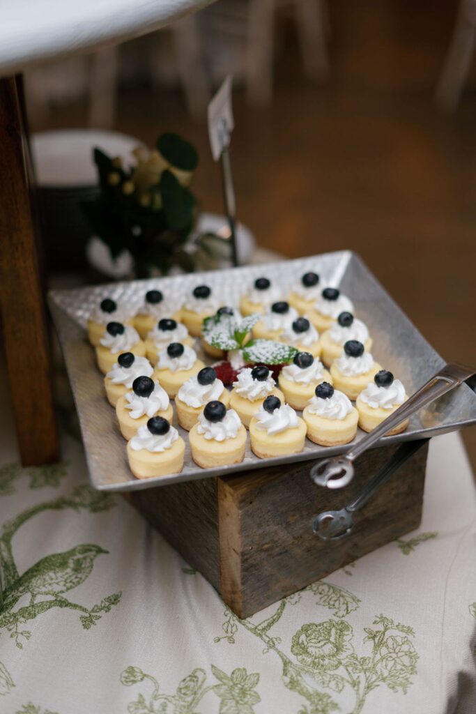 A metal tray filled with small, round desserts topped with whipped cream and a blueberry, displayed on a rustic wooden box at a Chastain Horse Park rehearsal dinner and garnished with mint. Metal tongs rest on the tray.