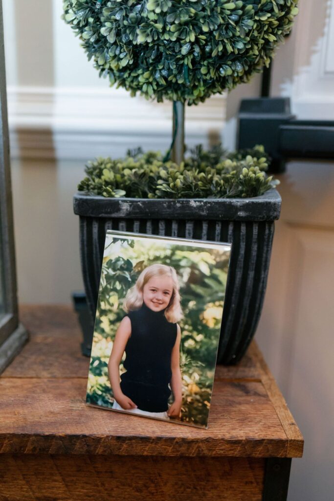 A framed photo of a young girl with blonde hair and a sleeveless black top is displayed on a wooden surface in front of a potted green plant at the Chastain Horse Park rehearsal dinner.